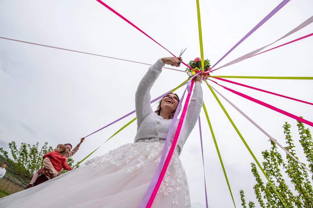 Photographie de Mariage de Ambre et Léo. Jeux du bouquet, ou la mariée lève le bouquet en l'air et doit couper une à une les ficelles de couleur ( rose, violet, vert ), la gagnante remporte le bouquet ! Photo prise en contreplongée, la mariée rit aux éclats dans sa belle robe blanche. Photographie prise par un photographe de mariage professionnel