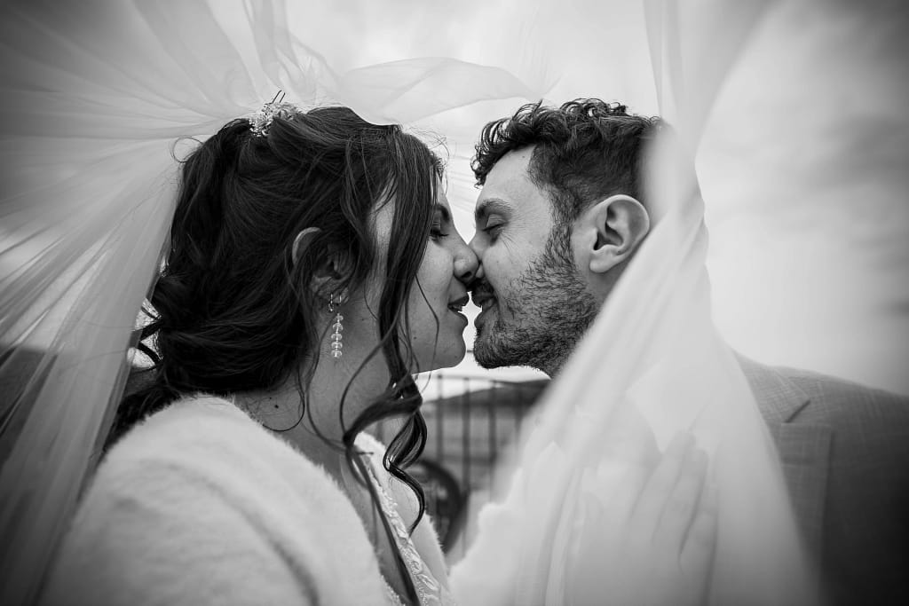 photographie de mariage Ambre et Léo. Photo de couple en noir et blanc. Les deux mariés s'embrasse sour le voile, photo prise au 24 mm avec un effet de proximité et du voile qui fait un ligne directrice vers les deus amoureux. Photographie prise par un photographe de mariage professionnel