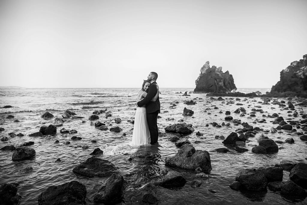 Photographie de mariage, Mathilde et Guillaume, photo à la plage de la grande conque. Ils sont au millieu des rochers dans l'eau et se fond un calin, on peut voir en arrière plan le grand rocher et les petits rochers en premier plan. Photo en noir et blanc. Photographie prise par un photographe de mariage professionnel