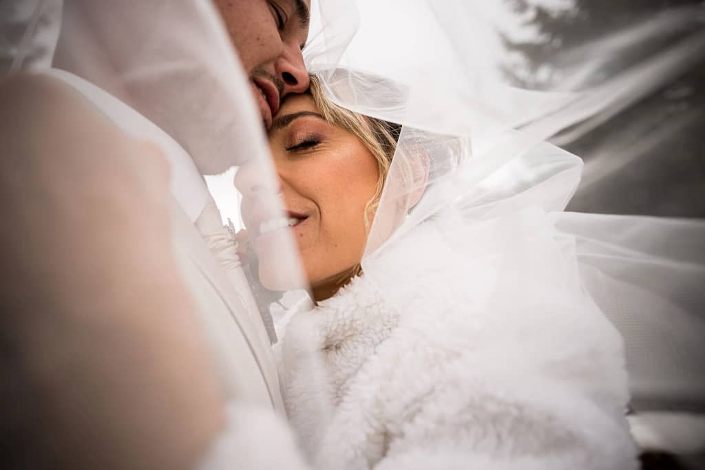 Photo de Mariage de Priscila et Steven. Photo en couleur,proche des deux mariés sous le voile. Photo faite au 24 mm, tête contre tête avec en premier plan la main de Priscilla et le voile qui les entoure. Photographie prise par un photographe de mariage professionnel