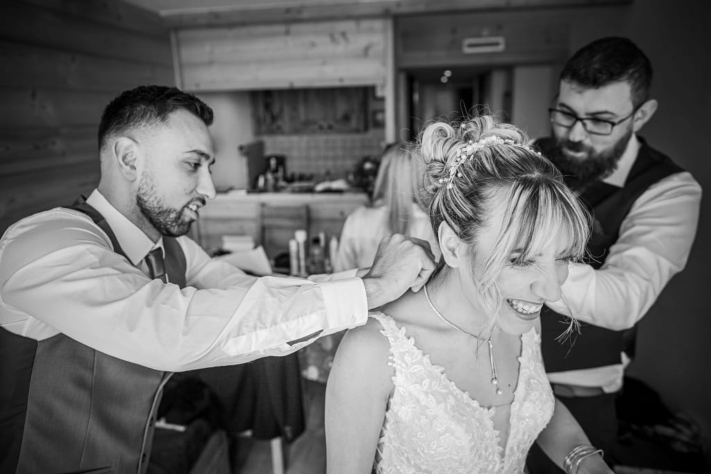 Photo du mariage de priscila et Steven, durant l'habillage de la mariée. Elle rit au éclat pendant que ses deux témoins sont frère et son cousins lui attache son collier. photo prise en plongée et en noir et blanc Photographie prise par un photographe de mariage professionnel