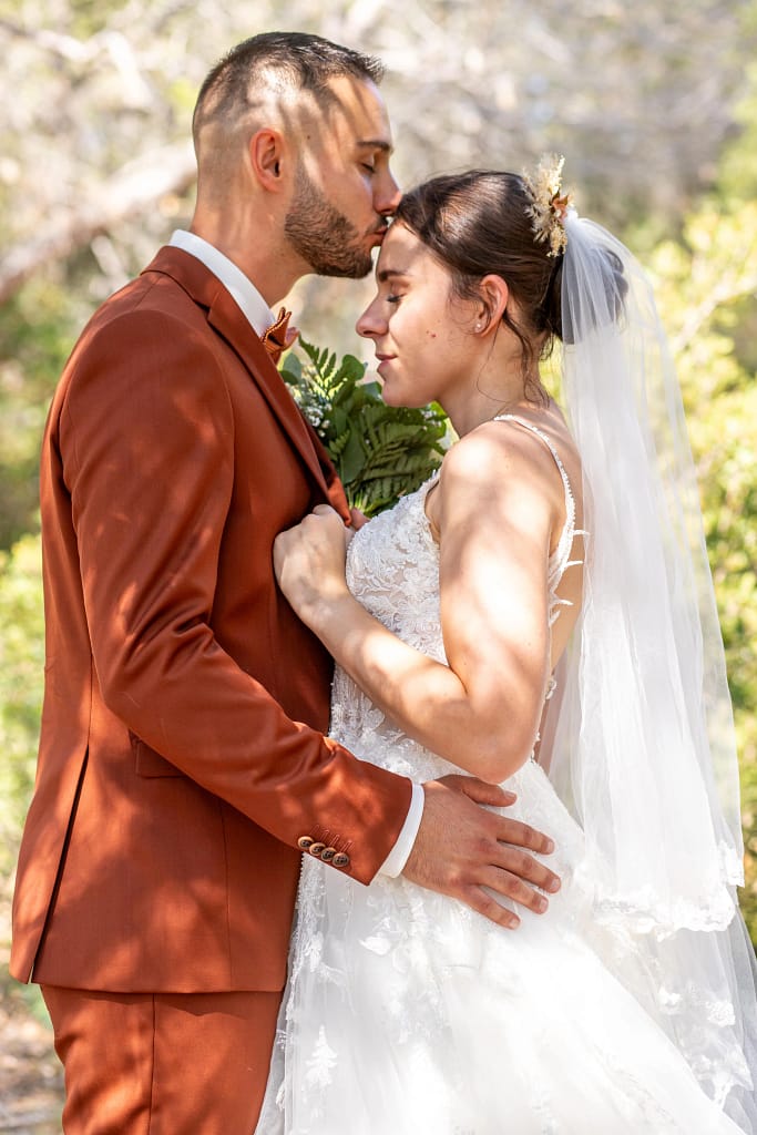 Photographie de mariage de Mathilde et Kévin. Kévin en tenue de cérémonie marron et Mathilde avec sa belle robe blanche sont face à face en extérieur, durant la séance de couple. Kévin l'embrasse avec amour sur le front tandis que Mathilde profite du moment les yeux fermés perdu dans ses pensés. Photographie prise par un photographe de mariage professionnel