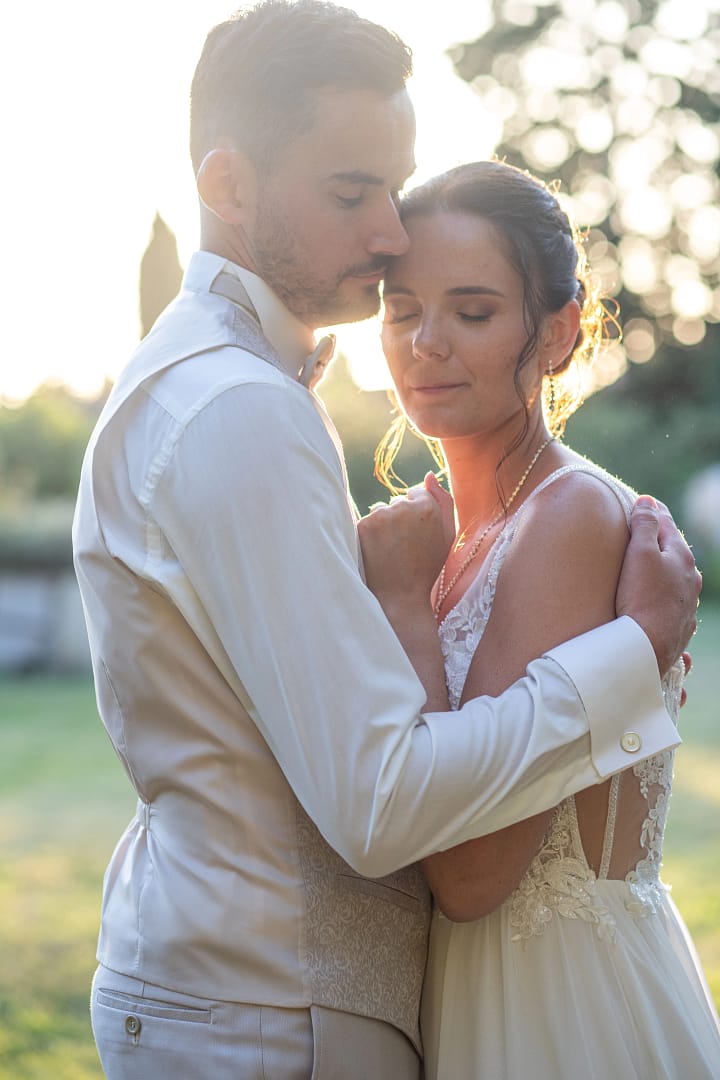 Photographie de mariage de Manon et Adrien en couleur à contrejour. Adrien tiens avec tendresse Manon dans ses bras et les deux amoureux profite du moment de calme avant l'entrée en salle. Photographie prise par un photographe de mariage professionnel