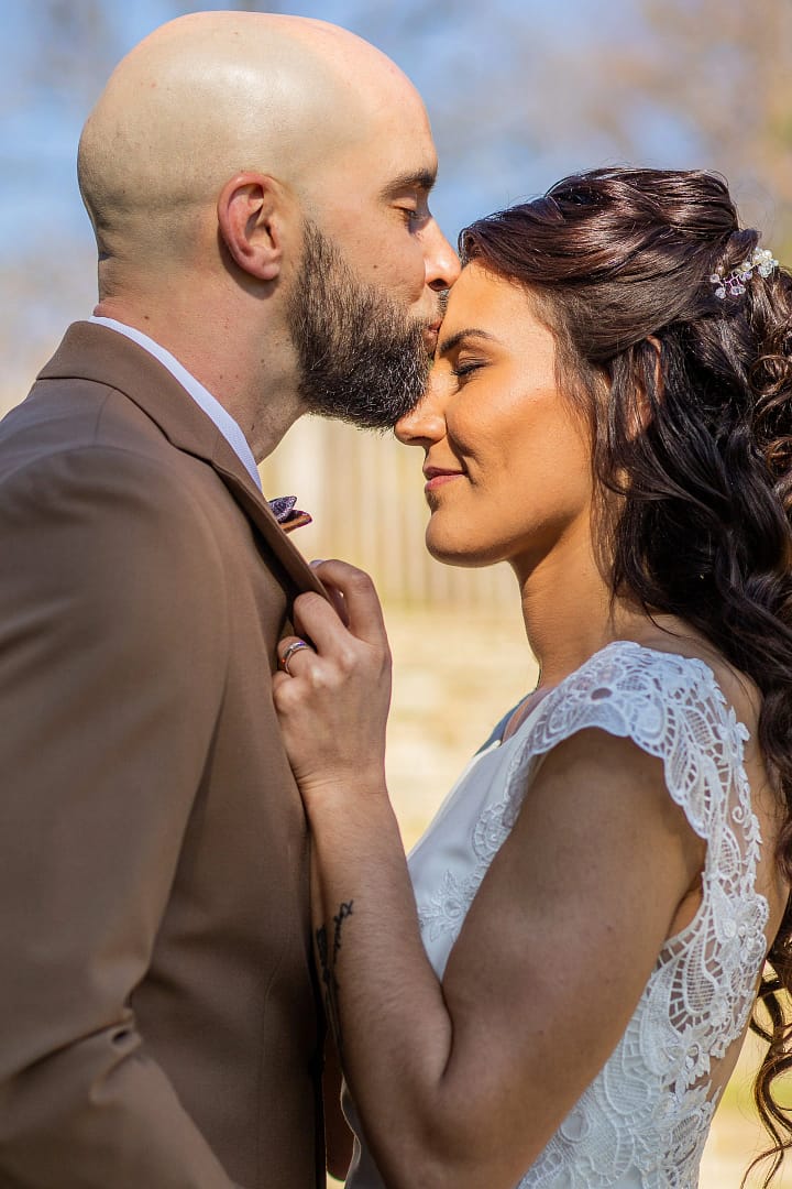 Mariage de Naomi et Romain. Romain dans son costume marron et Naomi dans sa robe blanche. Romain fait un superbe bisou sur le front de Naomi tandis qu'elle l'attrape par le col pour être encore plus proche de lui. Photo prise avec un cadrage vraiment serré. Photographie prise par un photographe de mariage professionnel