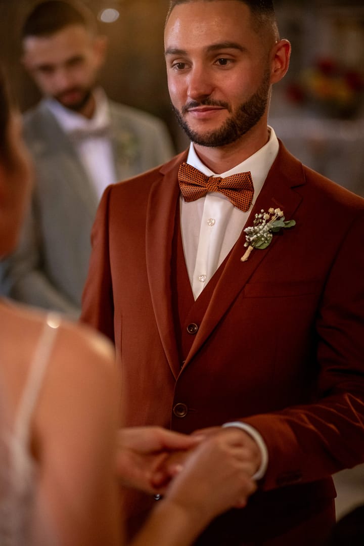 Photographie de mariage de Mathilde et Kévin. Kévin en tenue de cérémonie marron et Mathilde avec sa belle robe blanche. Ils sont debout dans l'église et Mathilde passe l'alliance au doigt de Kévin, tandis que lui la regarde avec amour . Photo en couleur. Photographie prise par un photographe de mariage professionnel