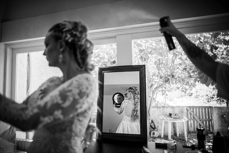 Photographie de mariage de Audrey et John. Photo en noir et blanc des prépartifs de la mariée. Elle est dos à un grand miroir, elle se regarde dans un petit miroir et la photo est faite pour que l'on voit son dos et son visage du petit miroir, dans le grand miroir tandis que la coiffeuse termine la coiffure à la laque. Photographie prise par un photographe de mariage professionnel