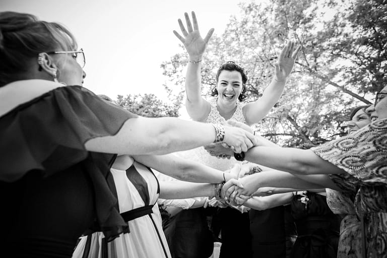 Photo de Mariage de Elisa et Alexis, la mariée se fait envoyer en l'air par tout les invités. Elle a les bras en avant tel superman et elle rit au éclats. Photo en noir et blanc. Photographie prise par un photographe de mariage professionnel