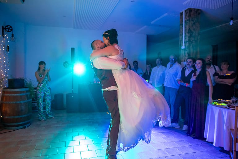 Photographie de mariage Élisa et Alexis. Première danse des deux amoureux du jour. Sur l'image il porte la mariée, elle se retrouve plus haut que lui et l'embrasse tendrement, avec le spot bleu du DJ en fond qui éclaire magnifiquement les mariés. Photographie prise par un photographe de mariage professionnel