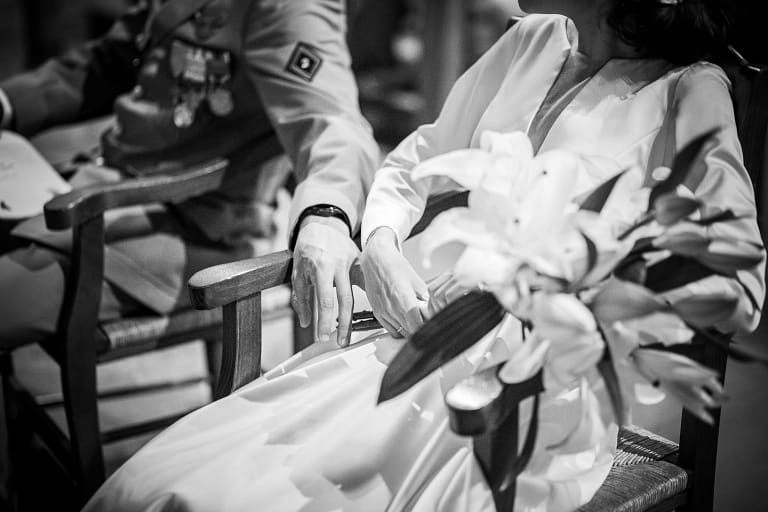 Photographie de mariage de Mahaut et Bryan. Bryan en tenue de cérémonie militaire et Mahaut avec sa belle robe blanche. Ils sont assis tout les deux à l'église et Bryan tant la main avec tendresse contre celle de Mahaut. Photo en noir et blanc Pas de visage que le buste est visible. Photographie prise par un photographe de mariage professionnel