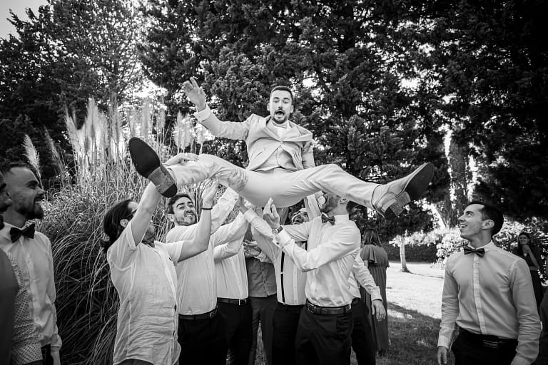 Photographie de mariage de Manon et Adrien en noir et blanc. Adrien se fait ne lancer en l'air pas un groupe de garçon et on voit sur son visage la frayeur du moment ou il est en plein vole. Photographie prise par un photographe de mariage professionnel