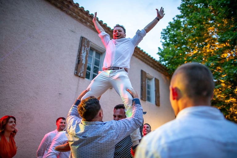 Photo de Mariage de Maxime et Muge en contreplongée . Un invité qui est projeter en l'air par deux autres invit&s, il le tiennent par les jambes et lui à les bras lever vers le ciel. Il porte une chemise blanche et un pantalon beige. Photographie prise par un photographe de mariage professionnel